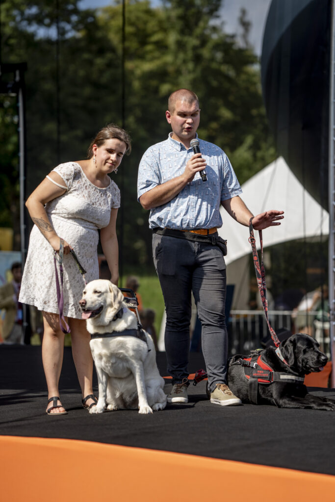 dwie osoby niewidome stoją na scenie z psami przewodnikami
